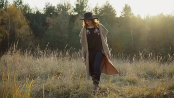 Wanita berambut merah dengan topi hitam dan mengenakan mantel berjalan di antara padang rumput — Stok Video