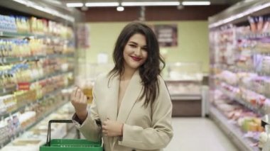 Elinde sepeti olan bir kadın süpermarket rafları arasındaki kameraya gülüyor.