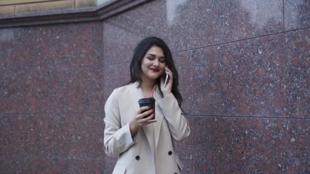 Jovencita alegre habla por teléfono con una sonrisa y bebe al aire libre — Vídeos de Stock