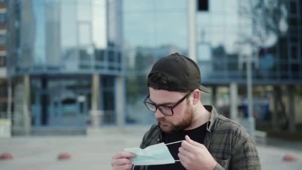 El joven con gafas y gorra lleva máscara médica al aire libre en la ciudad — Vídeos de Stock