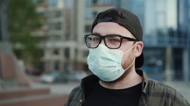 Portrait of young man in glasses, cap and medical mask blinks his eyes outdoors — Stock Video