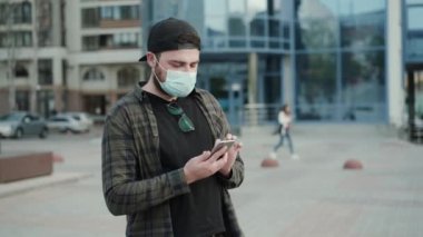 Tıbbi maske takmış genç bir adam şehirde sokakta akıllı telefon kullanıyor.