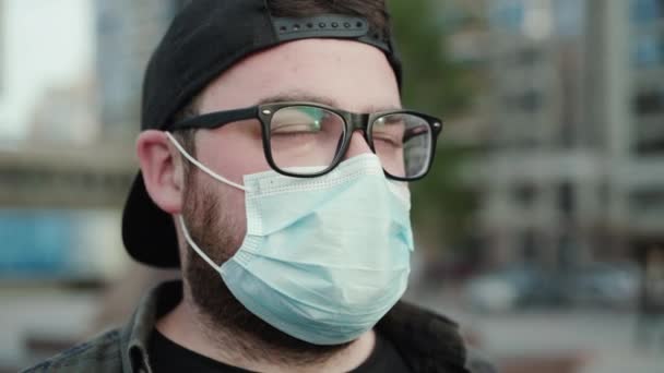 Retrato de hombre joven en gafas, gorra y máscara médica mira a un lado pensativamente — Vídeos de Stock