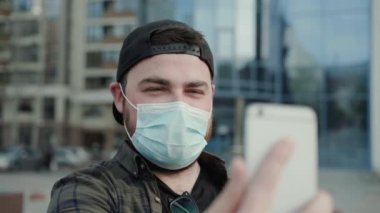 Tıbbi maskeli mutlu adam internette sohbet ediyor ve akıllı telefonlara el sallıyor.