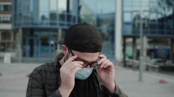 Joven hombre en máscara médica ponerse gafas y mirando a la cámara al aire libre — Vídeo de stock