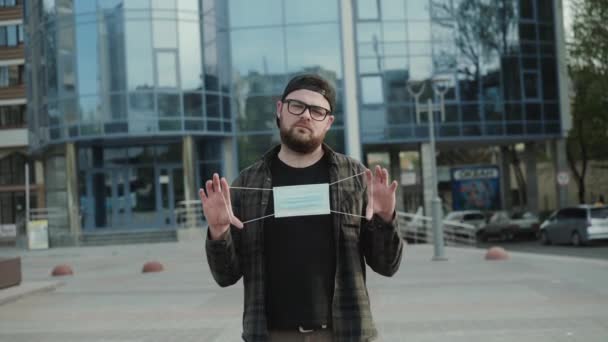 En ung man med glasögon sträcker medicinsk mask framför kameran på torget — Stockvideo