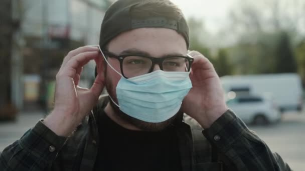 Young man wears protective medical mask corrects it and looks at camera outdoors — Stock Video