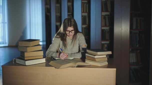 Una giovane ragazza annota diligentemente le informazioni dai libri in biblioteca tardi. — Video Stock