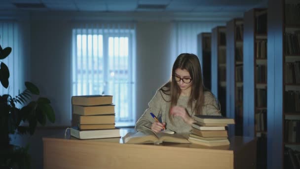 Vacker ung flicka med glasögon studerar i biblioteket, anteckningar från böcker — Stockvideo