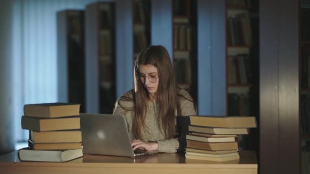 Menina jovem em óculos trabalhando duro com laptop e livros na mesa na biblioteca — Vídeo de Stock