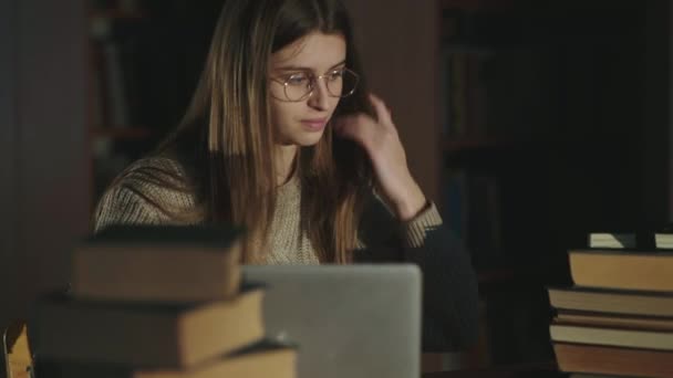 Bela estudante usa óculos e notas de laptop em uma biblioteca no final do tempo — Vídeo de Stock