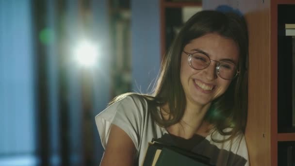 Menina de óculos, com livros nas mãos e inclinando-se na estante, ri na biblioteca — Vídeo de Stock
