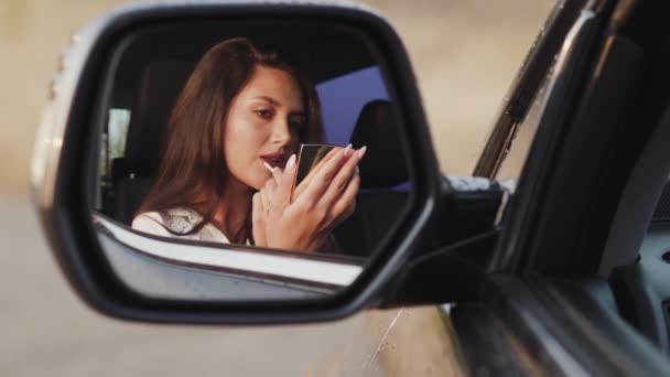 Отражение женщины в зеркале заднего вида автомобиля, которая смотрит на губы и улыбается — стоковое видео