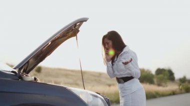 Endişeli iş kadını şehir dışında açık araba kaputunda telefonla konuşuyor.