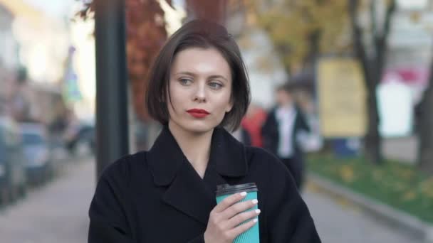 Гламурная леди в черном пальто с чашкой кофе стоит в позах на улице — стоковое видео