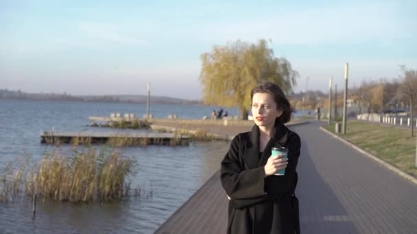 Жінка з чашкою напою повільно ходить біля міського озера і думає — стокове відео