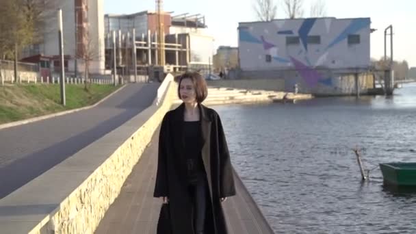 Красивая женщина с извилистыми волосами гуляет в пальто на городском озере — стоковое видео
