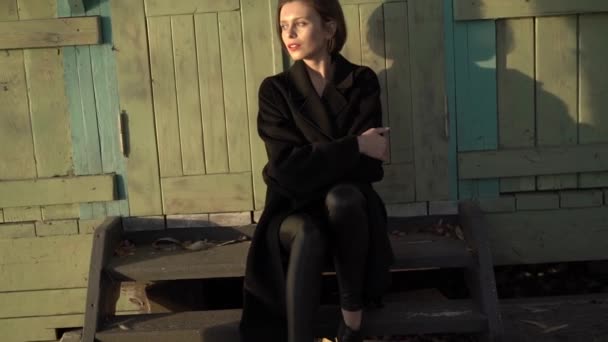 Modelo feliz envuelto en elegante abrigo negro relajarse en viejas escaleras de madera en la puesta del sol — Vídeos de Stock