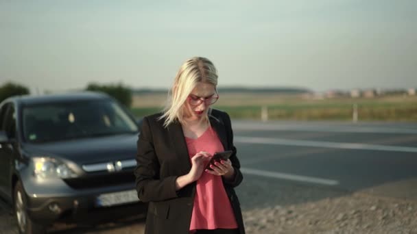 Bonita rubia con gafas escribiendo en un smartphone cerca de un coche roto cerca de la autopista — Vídeos de Stock
