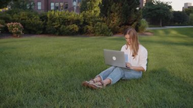Laptop 'ta çalışan genç bir kadın, şehir parkında çimenlikte oturuyor, serbest çalışan bir kadın, yaz parkında uzaktan çalışıyor. Dışarıda bir öğrenci var.