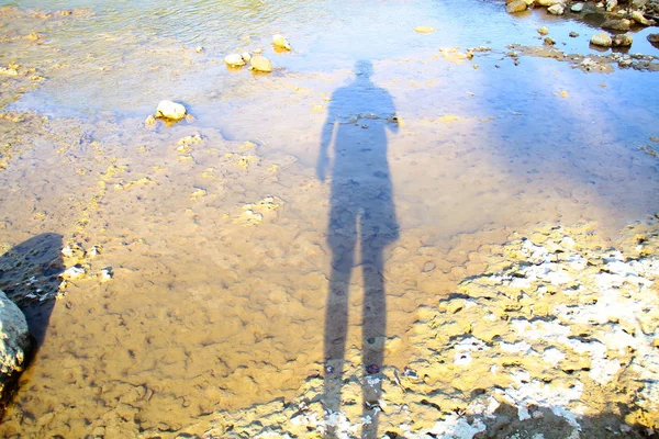 man\'s shadow reflected in the river on the shoreline on a bright day