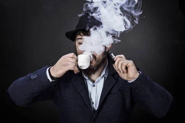 有胡子的家伙在帽子里喝咖啡 在黑色背景上吸烟电子雪茄 — 图库照片