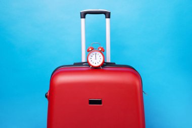 Mavi arka planda çalar saati olan bir seyahat çantası. Yolculuk zamanı. Yolculuk konsepti. Kırmızı valizli geri alarm saati