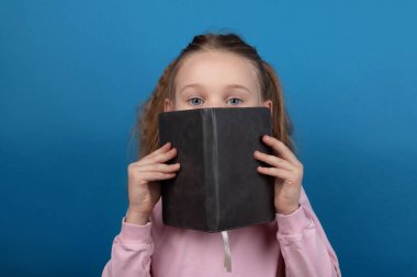 Pembe kazaklı genç kız, mavi arka planda izole edilmiş bir kitap okuyor. Kapağa bakan çocuk, eğitim, merak ve öğrenme kavramı.