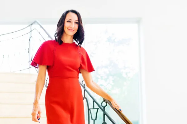 A girl in a red dress goes down the stairs. A young brunette is walking down the stairs. A woman in a red dress greets guests with a smile.