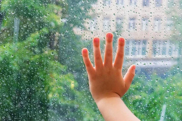 Children\'s hand on a wet glass with drops. The child outside the window looks at the autumn rain