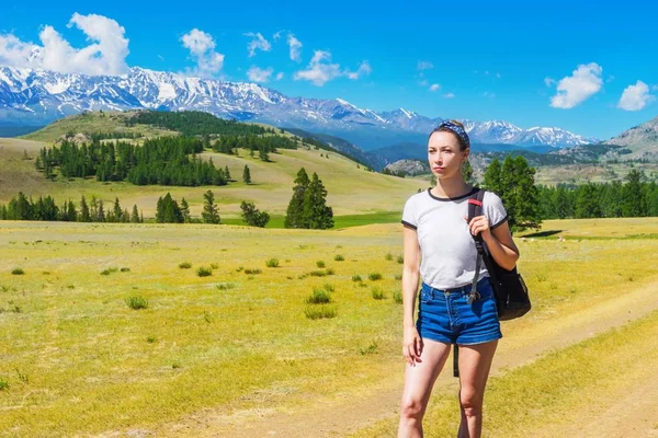 A girl with a backpack is walking along a path in a mountain valley. Tourist at a snowy ridge on a summer day