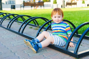Oğlan kaldırımda oturuyor. Şort ve tişört içinde küçük bir çocuk yolda oturdu. Bisiklet park yeri. Yaz gecesi.