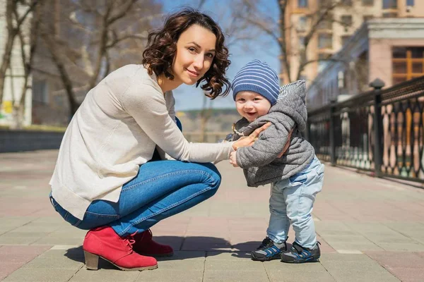 Mom teaches baby to walk on the street. Mom supports her son with her hands, teaches him to stand. A little boy learns to walk.