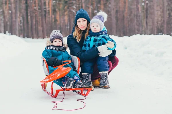 Mom with two children in a winter park. Family walk. A girl and two sons are walking along a snowy road. The boy is sledding next to his mother and brother.