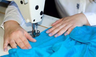 Terzi. Bir kadın dikiş makinesine mavi kumaş karalıyor..