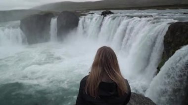 İzlanda'daki güçlü şelale bir kayaya arıyorum ve yalnız oturan genç dalgın kadın arkadan görünüm.