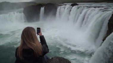 Kadın turist tarafından şelale Skogafoss İzlanda alarak fotoğraf ile akıllı telefon açık. Kız ziyaret ünlü turistik mekanlara ve İzlanda doğa manzara simge yapılar