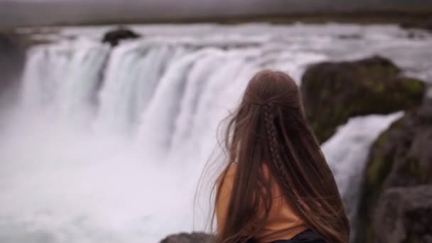 Back view of young pensive woman sitting on a rock alone and looking on powerful waterfall in Iceland. — Stock Video