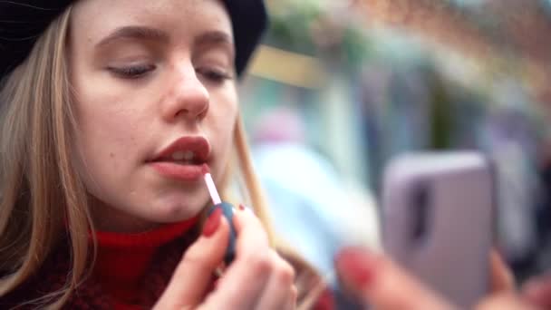 Jeune femme élégante attrayante peint ses lèvres avec du rouge à lèvres en regardant le reflet dans le téléphone à l'extérieur . — Video