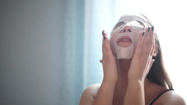 Jovem mulher fazendo a folha de máscara facial com máscara purificante em seu rosto no fundo branco — Vídeo de Stock