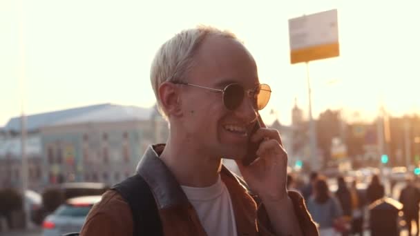 Молодой блондин держит мобильный телефон, использует смартфон, делает звонок, разговаривает по телефону, стоит на солнечной улице с транспортным движением на заднем плане — стоковое видео