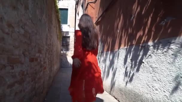 Una giovane ragazza con un vestito rosso corre lungo una strada stretta e si gira. Venezia, Italia . — Video Stock