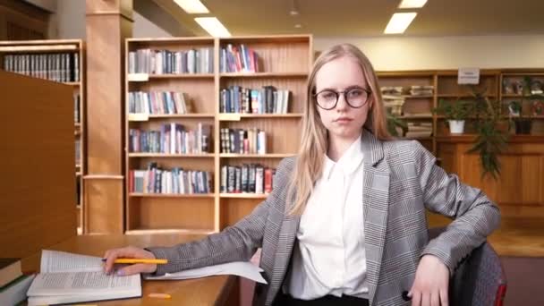 微笑美丽的学生站在图书馆的肖像。背景里的书柜书架 — 图库视频影像