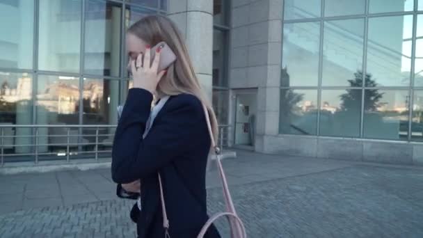 Vista desde un lado de una joven mujer de negocios caminando cerca de la pared, hablando por teléfono — Vídeos de Stock
