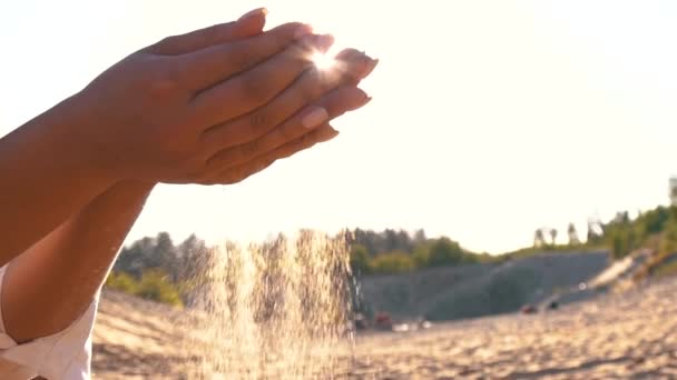Närbild av kvinna hälla sand rinner genom fingrarna slow motion på stranden med sol flare och blå himmel. Vacker scen av ung dam leker med sand på soluppgång eller solnedgång. — Stockvideo
