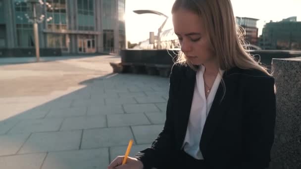 Kaukasische jonge zakenvrouw in wit overhemd zit op een bankje op straat en werkt. Het meisje schrijft in een notebook — Stockvideo