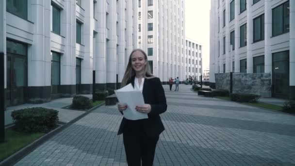 Young business women or student black suit look at paper documents, smile and throw them in to the air. Modern, contemporary glass building on background — Stock Video