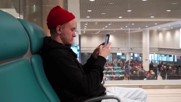 O jovem está tocando o número no celular ao lado da placa de informações no aeroporto. Homem atraente em chapéu vermelho falando no telefone celular . — Vídeo de Stock