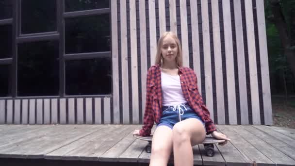Хипстерская девушка сидит на скейтборде перед деревянной стеной — стоковое видео