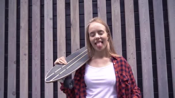 L'adolescente montre une langue et rit. Une jeune belle femme, avec un sourire blanc neige, pose sur le fond d'un mur de bois et tient une planche à roulettes sur ses épaules . — Video